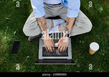 Ein reifer Mann mit Tattoos sitzt draußen auf dem Gras und tippt auf seinem Laptop. Stockfoto