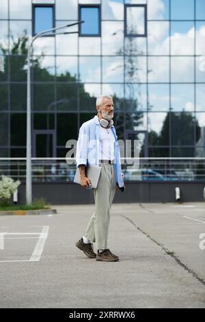 Ein reifer schwuler Mann mit Tattoos und grauem Bart geht mit einem Laptop durch einen Parkplatz vor einem Glasgebäude. Stockfoto