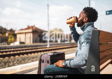 Ein Mann mit Koffer trinkt gern Kaffee, während er auf einer Bank am Bahnhof sitzt. Stockfoto