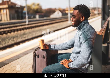 Glücklicher Mann mit Koffer trinkt Kaffee, während er auf einer Bank am Bahnhof sitzt. Stockfoto