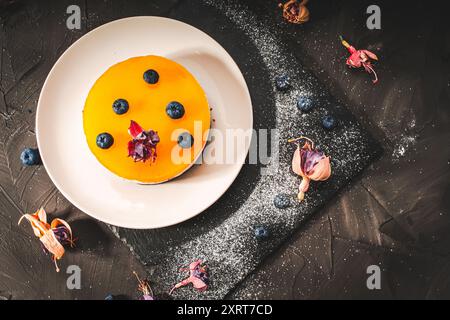 Dessert oder Kuchen mit Schichten Schokolade, Quark Creme, Mangogelee, dekoriert mit getrockneten Blumen auf einem weißen Teller auf schwarzem Hintergrund. Original Stockfoto