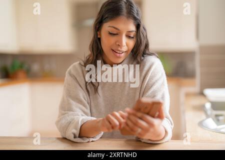 Glückliche junge indianerin, die ihr Handy benutzt, Lieblingslieder, Musik, soziale Medien, Hörbücher, Podcasts, ich rede, während ich in der Küche sitze Stockfoto
