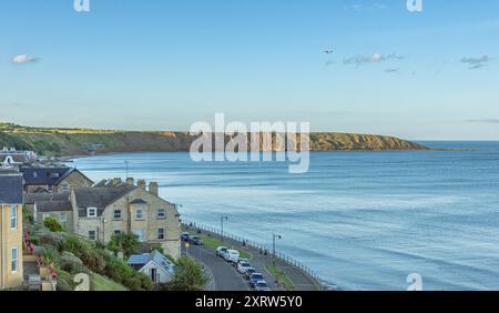Eine Bucht am Meer mit Häusern im Vordergrund und den Klippen einer Landzunge in der Ferne. Eine niedrige Abendsonne fängt Teile der Klippe. Stockfoto