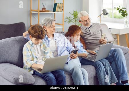 Glückliche Großeltern mit zwei Kindern, die zusammen mit Laptops und Mobiltelefonen auf dem Sofa sitzen. Stockfoto