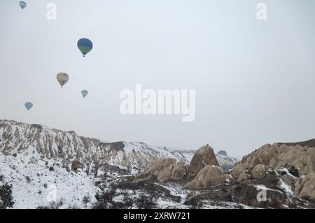 Heißluftballons, die über Kappadokiens schneebedeckter Landschaft schweben, zeigen felsiges Gelände und Feenkamine an einem ruhigen Wintermorgen Stockfoto