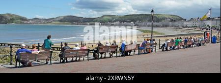 Rückansicht Erwachsene Touristen sitzen auf langen Sitzbänken breite Promenade Panoramablick auf Little Orme Llandudno Conwy Clwyd North Wales UK Stockfoto