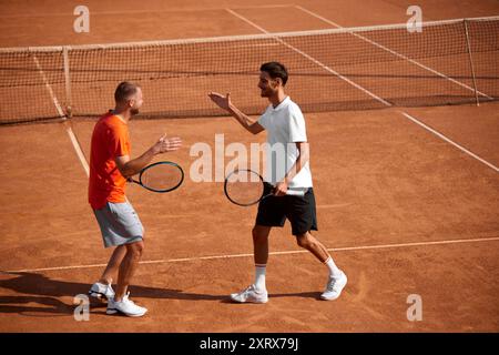 Zwei emotionale glückliche Männer, Tennisspieler in Bewegung auf dem Tonplatz, die sich gegenseitig unterstützen und Respekt zeigen, nach dem Golfspiel die Hand schütteln Stockfoto