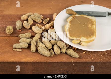 Erdnüsse in der Schale mit ein paar gebrochenen Schalen, die auf einem rauen Holztisch liegen, mit Erdnussbutter auf einer Brotscheibe auf weißem Teller Stockfoto