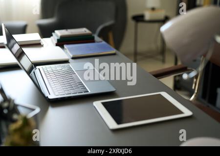 Organisierter Bürotisch mit Laptop, Tablet und Bücherstapel. Die Gegenstände sind ordentlich unter sanfter Innenbeleuchtung angeordnet und präsentieren sich sauber und produktiv Stockfoto