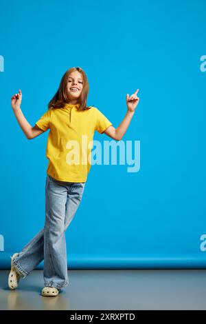 Das ganze Bild eines glücklichen jungen Mädchens in gelbem Poloshirt und blauer Jeans tanzt vor blauem Studiohintergrund Stockfoto