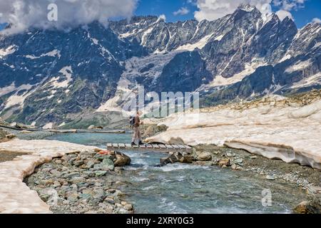 Eine Wandererin mit Rucksack überquert einen sintflutartigen Bach auf einer schmalen Holzbrücke zwischen Schneefeldern in den alpen nahe der matterhornlandschaft Stockfoto