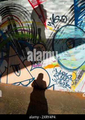 Graffiti und Schatten unter einer Brücke. Kontraste. Dieses Bild von Graffiti an der Themse in der Nähe von Oxford zeigt nicht den Verkehr, der Tag und Nacht auf der Brücke vorbeifließt und den verstopften Eastern Bypass über den Fluss trägt. Mein Schatten scheint auf den ersten Blick ziemlich gut in das Kunstwerk zu passen Sie fragen sich vielleicht, warum ich zwei Schatten habe. Einfach. Der untere ist ein direkter Schatten von der frühen Sonne, die unter der Brücke kriecht; der obere, gesprenkelte ist von der Sonne, die von der zerrissenen Oberfläche des Flusses reflektiert wird. Optik in Aktion... Stockfoto