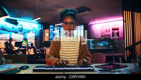 Porträt einer schwarzen Entwicklerin, die Code in einem Kreativbüro mit Neonlichtern schreibt. Junge Frau, die sich auf das Tippen auf der Tastatur und die Arbeit am Desktop-Computer konzentriert. Point of View des Bildschirmmonitors Stockfoto
