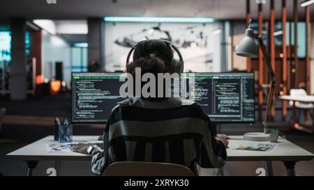 Creative Office: Der professionelle Schwarze Programmierer verwendet Kopfhörer, die auf einem Desktop-Computer arbeiten. Focus Coder, Software Engineer, Entwickelt App, Videospiel. Podcast hören, Musik. Rückansicht. Stockfoto