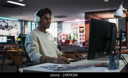 Porträt eines stilvollen Indianers, der in einem Firmenbüro am Desktop-Computer arbeitet. Junger männlicher Manager, der im Internet surft, online shoppt und Beiträge von Freunden und Kollegen aus sozialen Medien liest Stockfoto