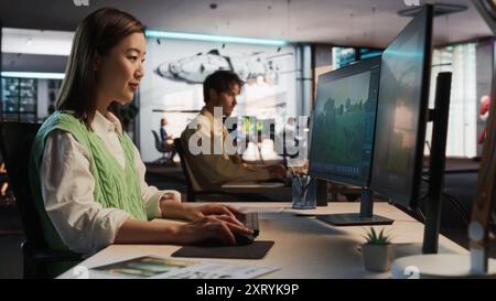 Asiatische Frau, die Desktop-Computer verwendet und in 3D-Modellierungssoftware Unique World und Charaktere für Survival Videospiel in diversen Büros entwirft. Weibliche Spieledesignerin, Die Immersives Gameplay Kreiert Stockfoto