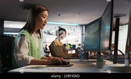 Asiatische Frau, die Desktop-Computer verwendet und in 3D-Modellierungssoftware Unique World und Charaktere für Survival Videospiel in diversen Büros entwirft. Spieleentwicklerin Für Ein Immersives Gameplay Stockfoto