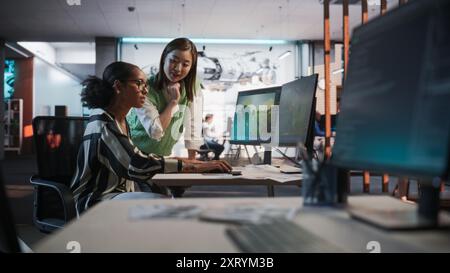 Black Female Game Designer verwendet Desktop-Computer mit 3D-Modellierungssoftware und spricht mit asiatischer weiblicher Projektmanagerin im Game Development Studio Office. Verschiedene Kollegen diskutieren über das neue Rollenspiel Stockfoto