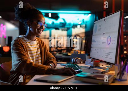Porträt einer glücklichen erfolgreichen schwarzen Geschäftsfrau, die am Abend einen Desktop-Computer in Creative Agency verwendet. Afroamerikanerin lächelt beim Prüfen von Daten- und Statistikkarten Stockfoto