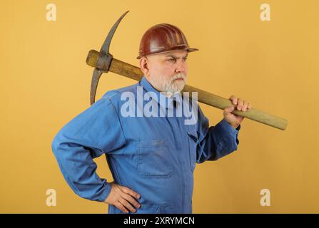 Schwerer Maurer im Schutzhelm mit Spitzhacke. Bärtiger Bauarbeiter im Overall und Schutzhelm mit Meißel. Handwerker, Bauunternehmer oder Bergarbeiter in Unif Stockfoto