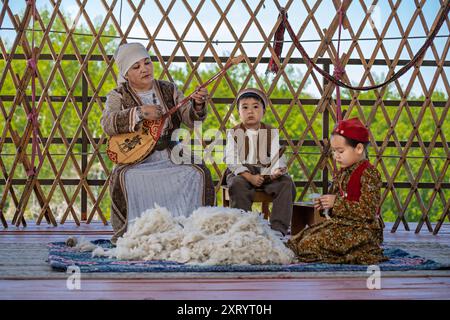 Nomadische Frau in traditioneller Kleidung spielt lokales Instrument, das als Dombra bekannt ist, während die Kinder sitzen und zuhören, in Almaty, Kasachstan. Stockfoto