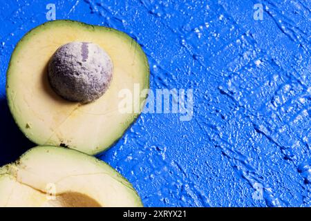 Zwei Teile einer Avocado auf der linken Seite des Bildes, eine Hälfte mit der Grube auf einem strukturierten blauen Hintergrund. Stockfoto