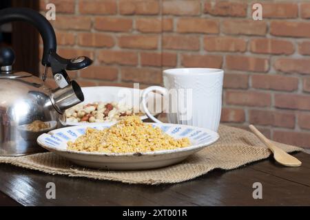 Hausgemachte Erdnussbrüchigkeit, eine Art Farofa, auf einem alten Teller mit Alterspuren, neben einem Wasserkocher und auf einer rustikalen Tischdecke. Stockfoto