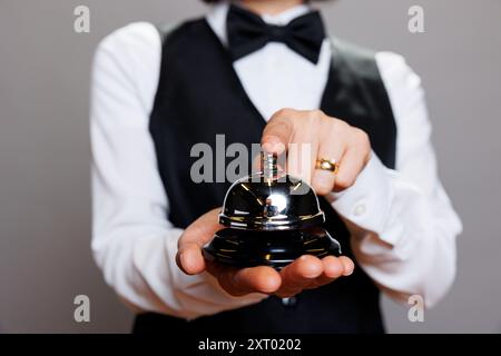 Die Kellnerin in Uniform hält die Essensglocke in den Händen und klingelt, während sie in der Nähe des Studios posiert. Catering-Service-Mitarbeiter drücken Auftragsbenachrichtigung mit Finger auf grauem Hintergrund Stockfoto