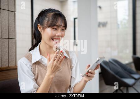Eine fröhliche, schöne asiatische Frau lacht und genießt es, sich mit ihren Freunden zu unterhalten oder Videos in sozialen Medien anzusehen, während sie sich drinnen ausruht. Stockfoto
