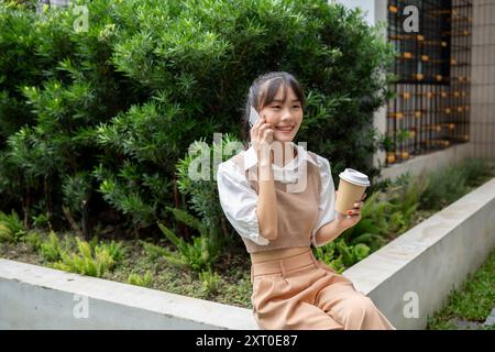 Eine attraktive, glückliche asiatische Frau trinkt Kaffee und spricht am Telefon während ihrer Mittagspause im Freien. Stockfoto