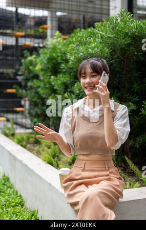 Eine schöne, glückliche junge asiatische Frau genießt eine Unterhaltung auf ihrem Telefon, während sie sich in einem Garten im Freien ausruht. Menschen und Wireless-Technologie sind verborgen Stockfoto