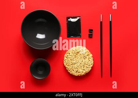 Rohe Instantnudeln mit leeren Schüsseln, Stäbchen und Gewürzen auf rotem Hintergrund Stockfoto