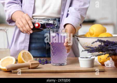 Junge Frau, die Lavendelsaft aus der Flasche in das Glas gießt, um Limonade am Tisch in der Küche zuzubereiten Stockfoto