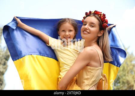 Niedliches kleines Mädchen und ihre Mutter mit ukrainischer Flagge und Blumenkranz vor blauem Himmel Stockfoto