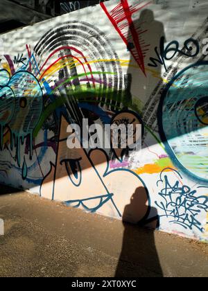 Kontraste. Dieses Bild von Graffiti an der Themse in der Nähe von Oxford zeigt nicht den Verkehr, der Tag und Nacht auf der Brücke vorbeifließt und den verstopften Eastern Bypass über den Fluss trägt. Mein Schatten scheint auf den ersten Blick ziemlich gut in das Kunstwerk zu passen Sie fragen sich vielleicht, warum ich zwei Schatten habe. Einfach. Der untere ist ein direkter Schatten von der frühen Sonne, die unter der Brücke kriecht; der obere, gesprenkelte ist von der Sonne, die von der zerrissenen Oberfläche des Flusses reflektiert wird. Optik in Aktion... Stockfoto