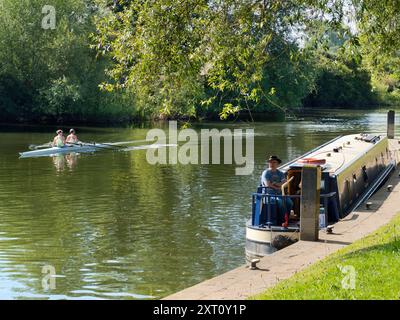 Die Gegend um Sandford Lock über der Themse ist ein beliebter Ort für Jogger, Spaziergänger, Hundeliebhaber und viele Leute, die die tollen Spaziergänge in dieser Gegend besuchen. Hier sehen wir zwei sehr unterschiedliche Wasserliebhaber - einen zwei-Mann-Schädel, der an einem eher entspannteren Hausboot vorbeirudert. Stockfoto