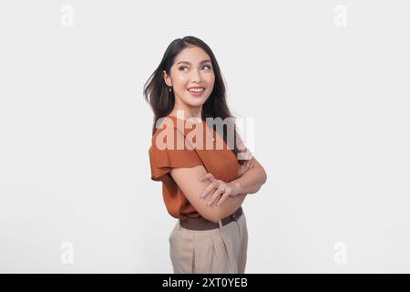 Charmante junge asiatische Frau in braunem T-Shirt, die oben mit überkreuzten Armen über isoliertem weißem Hintergrund schaut oder schaut. Stockfoto