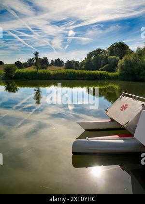 Das 1921 gegründete Radley Boathouse befindet sich an einem wunderschönen Teil der Themse in Oxfordshire und dient seit über einem Jahrhundert Radley College und lokalen Ruderfreunden. Wenn Sie auf dem Steg stehen und auf das Nordufer des Flusses blicken, sehen Sie einen isolierten und ziemlich schönen Baum, der Charakter und Aussehen im Laufe des Jahres zu verändern scheint. Hier sehen wir es an einem bewölkten Sommermorgen, mit den allgegenwärtigen Vergnügungskatamaranen des Bootshauses im Vordergrund. Das rote Malteserkreuz ist das Emblem des College Boot Club. Stockfoto