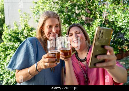 Zwei fröhliche Frauen im mittleren Erwachsenenalter lachen beim Selfie, wobei eine ein Smartphone hält und beide in einer üppigen Gartenumgebung ein Bierglas klirren Stockfoto