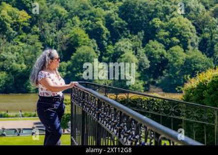 Ältere Erwachsene Frau im Profil auf Brücke mit Metallgeländer in Freyr Castle Gärten, Maas Fluss und grünen Laubbäumen in unscharfem Hintergrund, Stockfoto