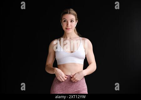 Eine fitte Frau in Activewear steht selbstbewusst vor einem schwarzen Hintergrund und ist auf ihre Trainingsroutine vorbereitet Stockfoto
