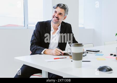 Ein umgänglicher Manager in schwarzem Anzug und weißem Hemd sitzt an seinem Schreibtisch, lächelt und macht Notizen, umgeben von wichtigen Arbeitsmitteln Stockfoto
