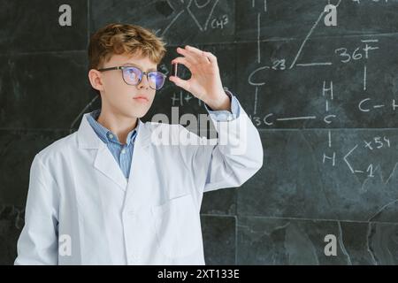 Fokussierter Jugendlicher in Brille, der auf das Reagenzglas blickt, während er vor der Tafel steht, mit schriftlichen chemischen Formeln auf dem Hintergrund im Klassenzimmer Stockfoto