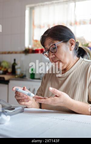 Eine ältere asiatische Frau mit Brille überprüft ihren Blutzuckerspiegel mit einem digitalen Glukometer, während sie an ihrem Küchentisch sitzt Stockfoto