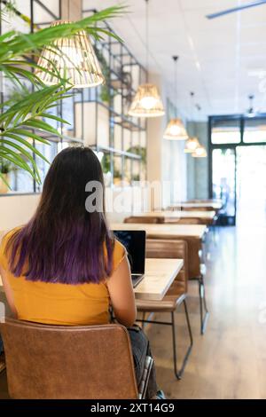 Rückansicht einer nicht erkennbaren jungen Unternehmerin, die am Tisch sitzt und Netbook durchstöbert, während sie am modernen Arbeitsplatz arbeitet Stockfoto