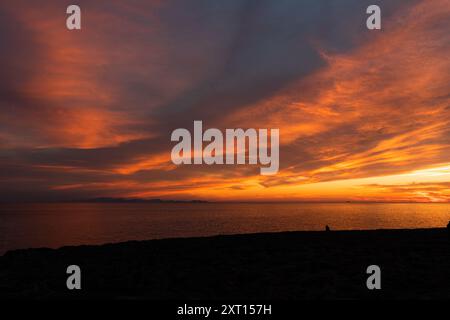 Spektakulärer Blick auf die Silhouette des Reisenden, der beim Sonnenuntergang den Ozean vom Strand aus unter einem farbenprächtigen, wolkigen Himmel betrachtet Stockfoto