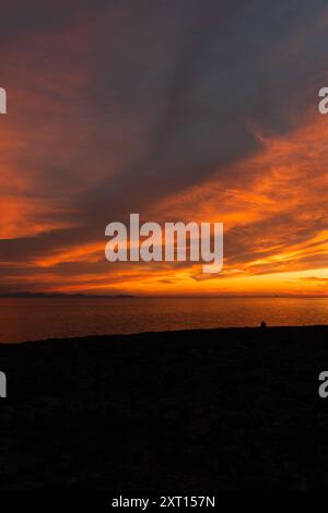 Spektakulärer Blick auf die Silhouette des Reisenden, der beim Sonnenuntergang den Ozean vom Strand aus unter einem farbenprächtigen, wolkigen Himmel betrachtet Stockfoto