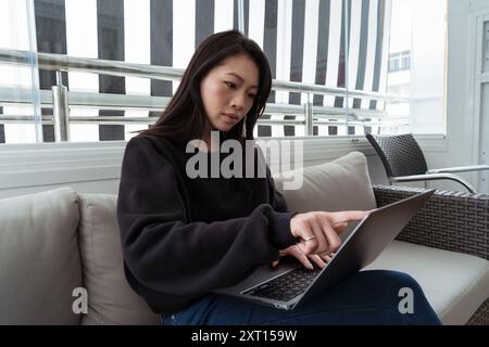 Eine fokussierte junge Frau benutzt einen Laptop, während sie bequem auf einer Couch sitzt, mit einem modernen gestreiften Vorhang im Hintergrund Stockfoto