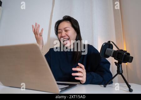 Eine fröhliche junge Frau streamt live über ein Smartphone, das auf einem Stativ montiert ist, und interagiert mit einem Laptop in einem zwanglosen Zuhause Stockfoto