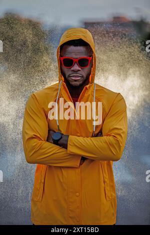 Ein selbstbewusster sportlicher Mann, der in einem auffälligen gelben Regenmantel mit einer auffälligen roten Sonnenbrille posiert, ist ein Beispiel für eine einzigartige Mischung aus Zweckmäßigkeit und Mode Stockfoto